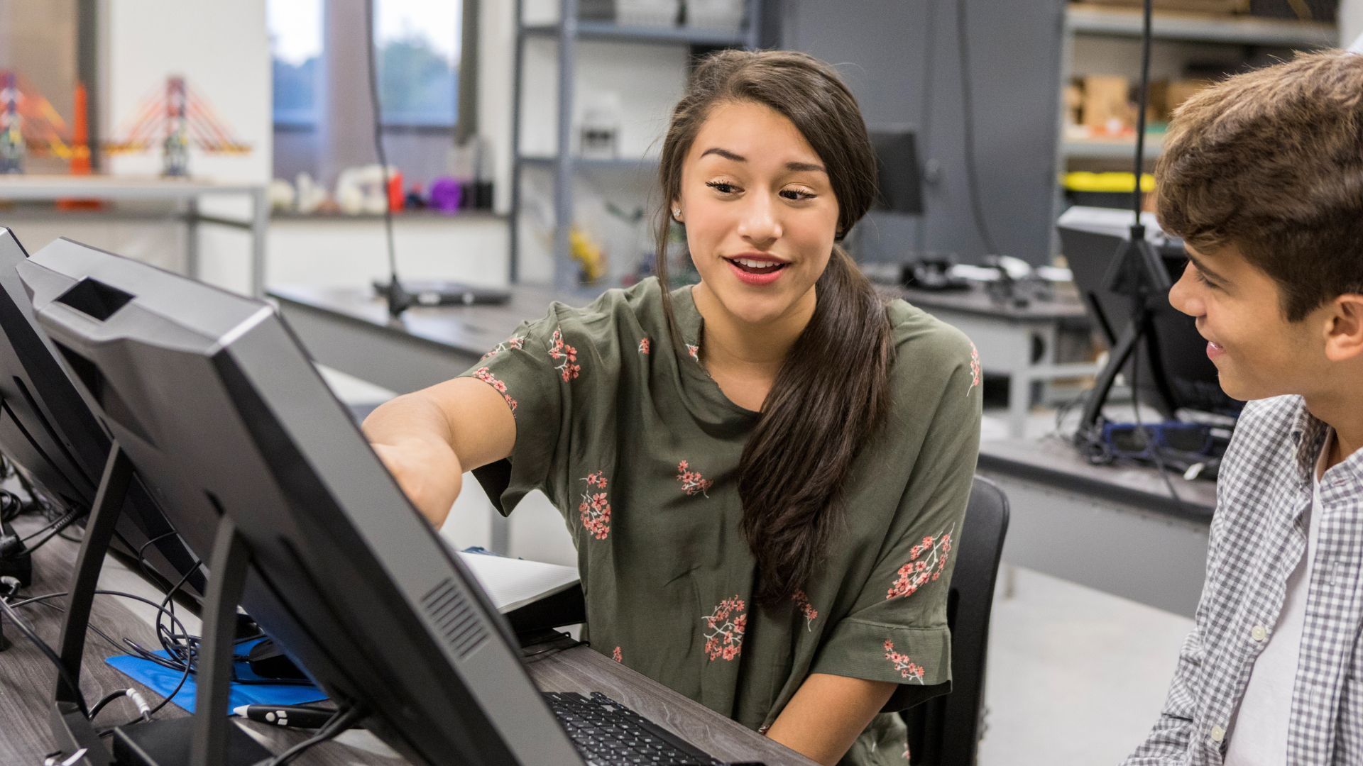 a imagem mostra uma adolescente mostrando algo a seu colega na tela do computador