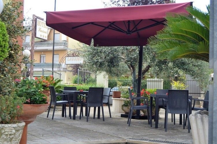 Caffetteria all'aperto con tavoli e sedie sotto un ombrellone rosso. Alberi e piante in vaso circondano il patio.