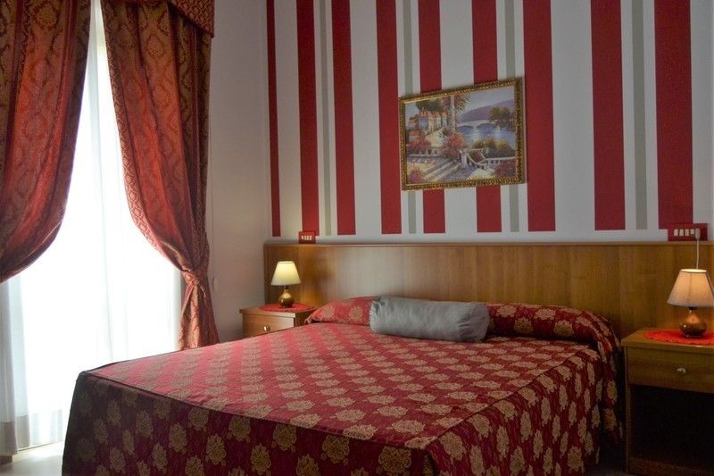 Camera d'albergo con carta da parati a righe rosse e bianche, letto matrimoniale, tende rosse e comodini.