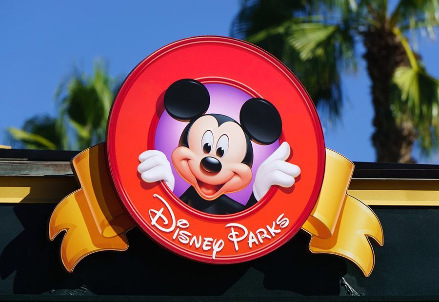 Mickey Mouse smiling in a red Disney Parks sign. Background has palm trees and blue sky.