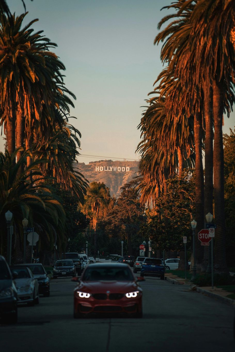 Red car driving towards the Hollywood sign, framed by palm trees at sunset.