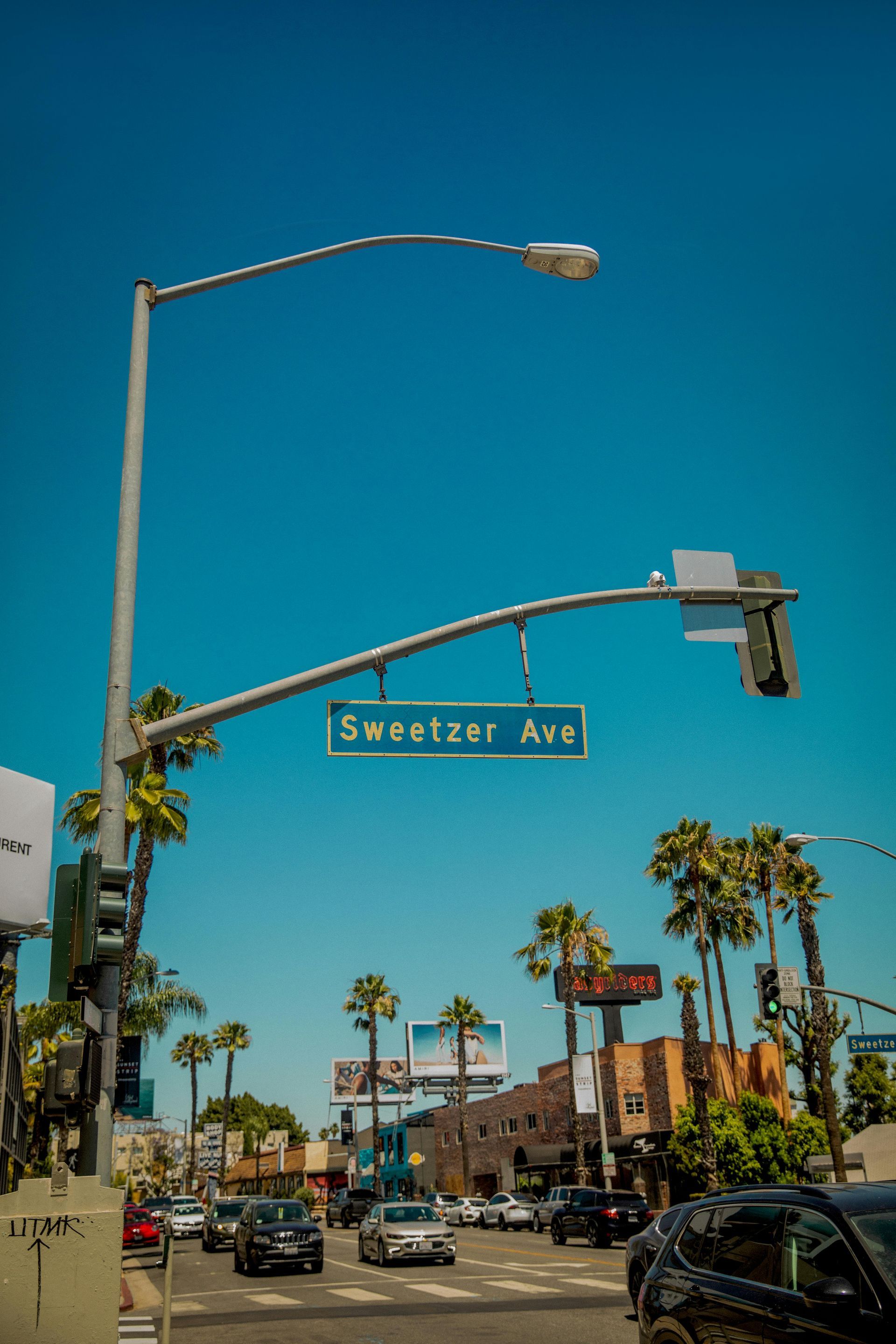 Street scene on Sweetzer Ave, Los Angeles, with palm trees, cars, and a clear blue sky.