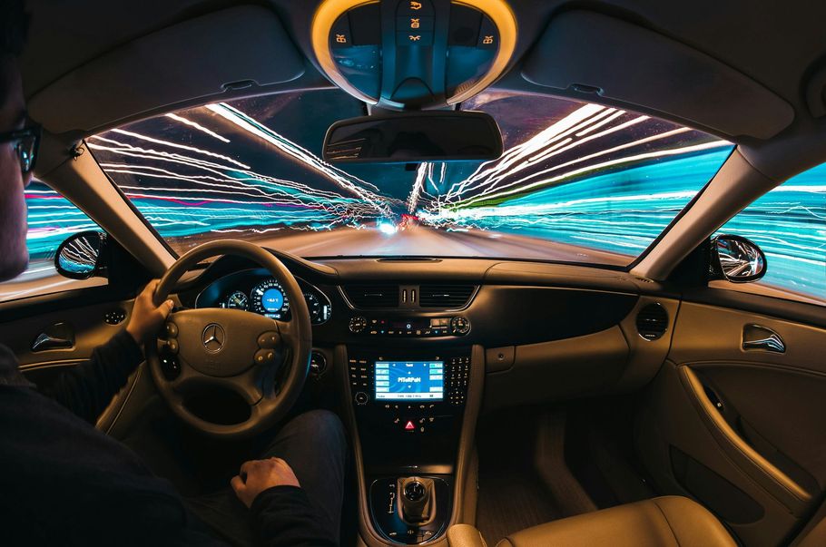 Inside a car driving on a road, blurred lights whizz past. Driver's hands on steering wheel.