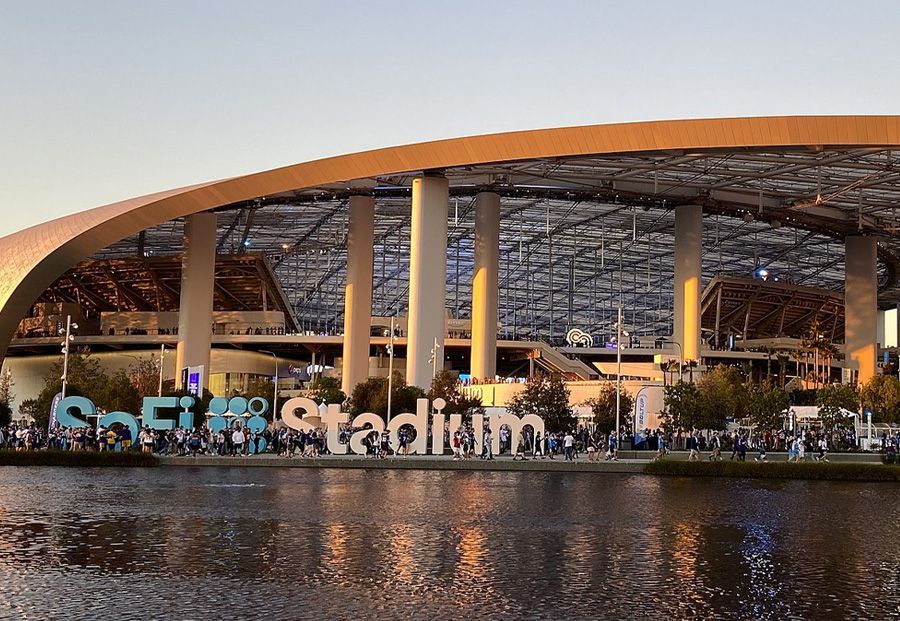 SoFi Stadium sign by a lake, people milling about. Building exterior in background.