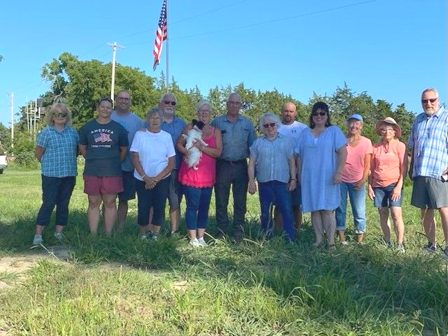Group of people standing outside under a blue sky with an American flag. Several people are smiling.