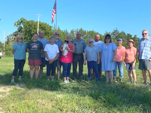 Group of people standing outside under a blue sky with an American flag. Several people are smiling.