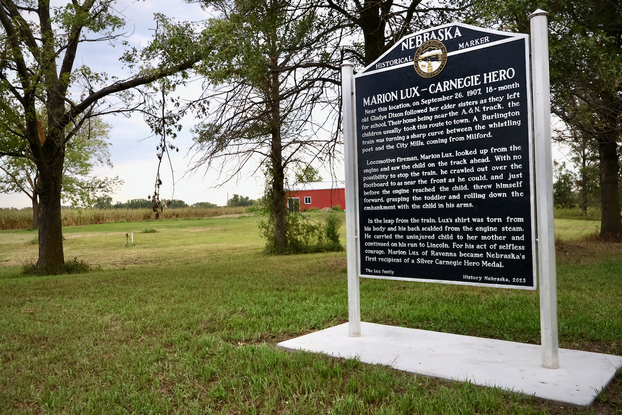 Historical marker in a grassy field, text on black background, trees, and a red building in the distance.