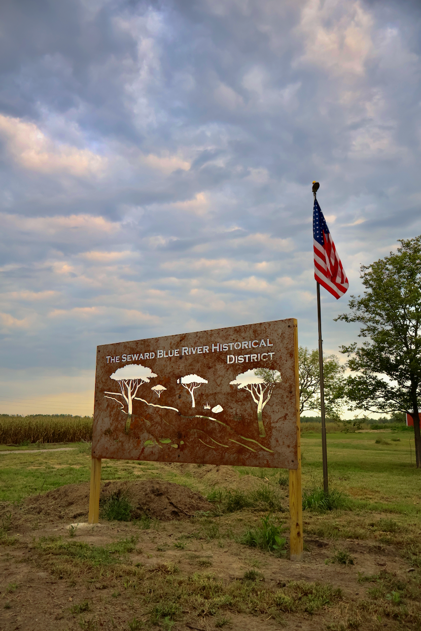 Sign with tree silhouettes, American flag, field, and cloudy sky.