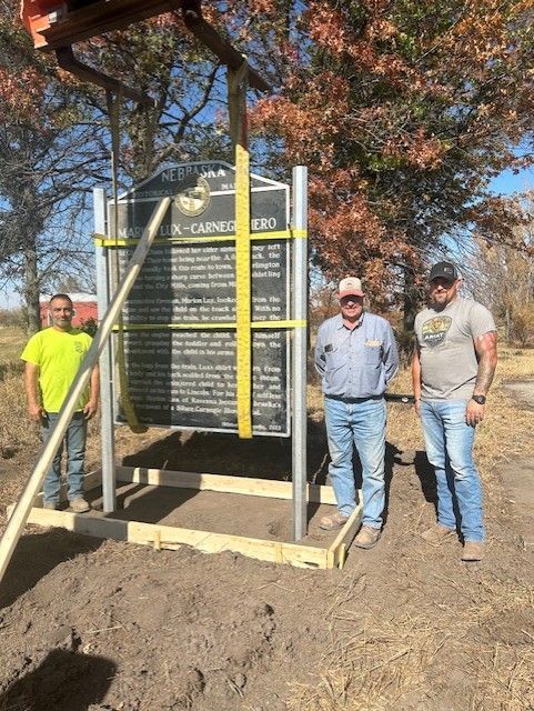 Three men installing a historical marker, held by straps and a crane, in a grassy outdoor setting.