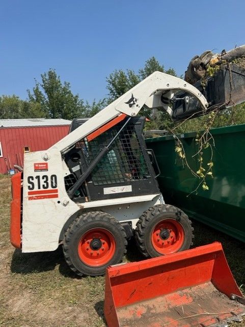White and orange Bobcat S130 skid-steer loader dumping debris into a green dumpster, outdoors.