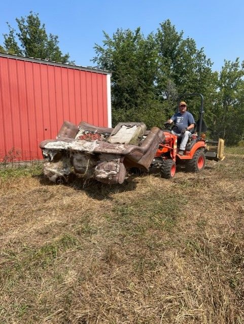 Man on a tractor hauling a dilapidated vehicle frame across a grassy field near a red barn.