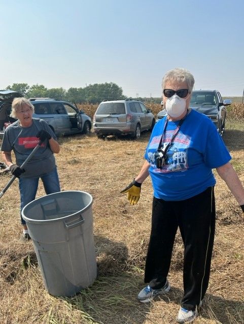 Two older women cleaning up trash in a field; one has a mask and sunglasses on; cars and dry grass are in the background.