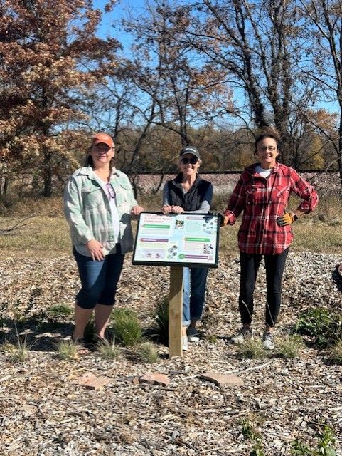 Three women stand near a sign in a natural setting. They are smiling.
