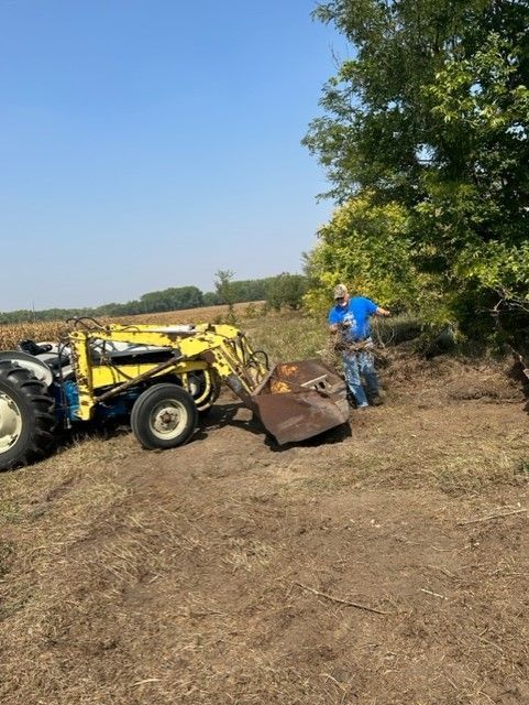 Man operating a yellow tractor with a loader outdoors; a blue sky and field are in the background.