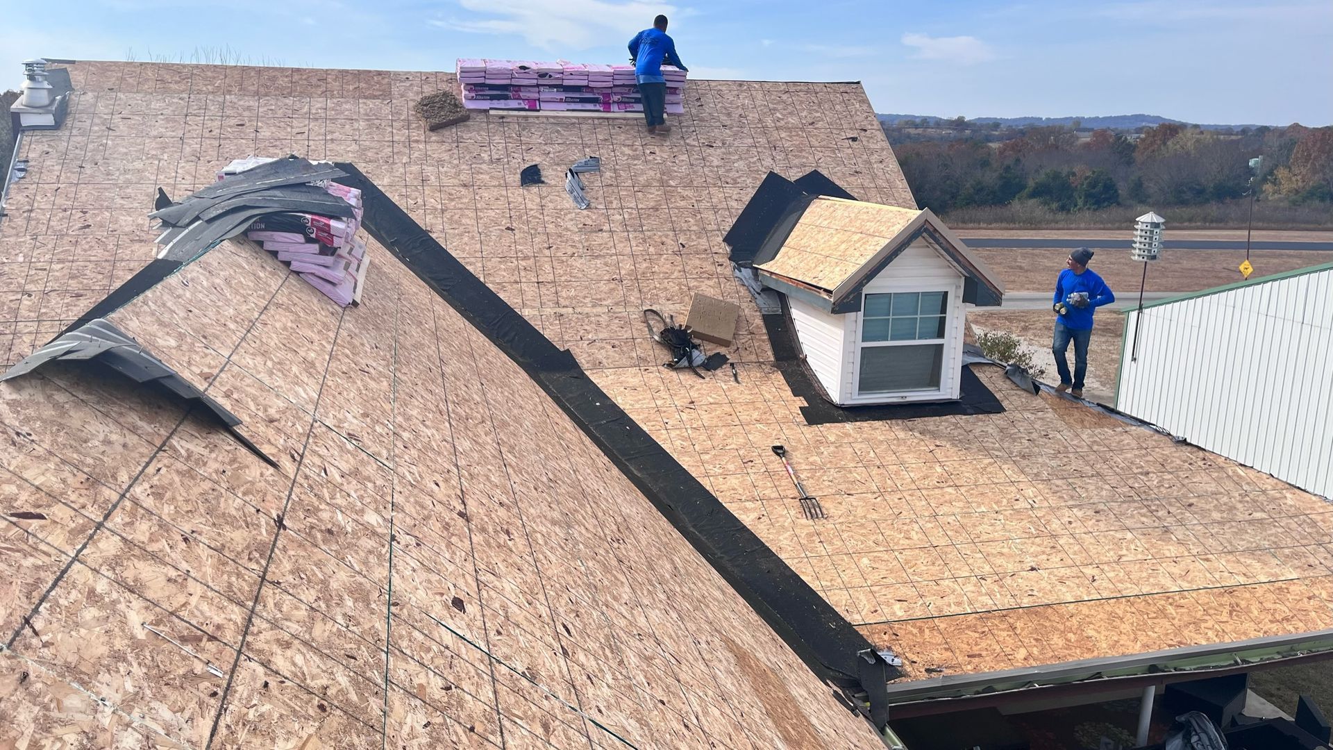 Roofers working on a house roof; visible plywood, insulation, and a small dormer.