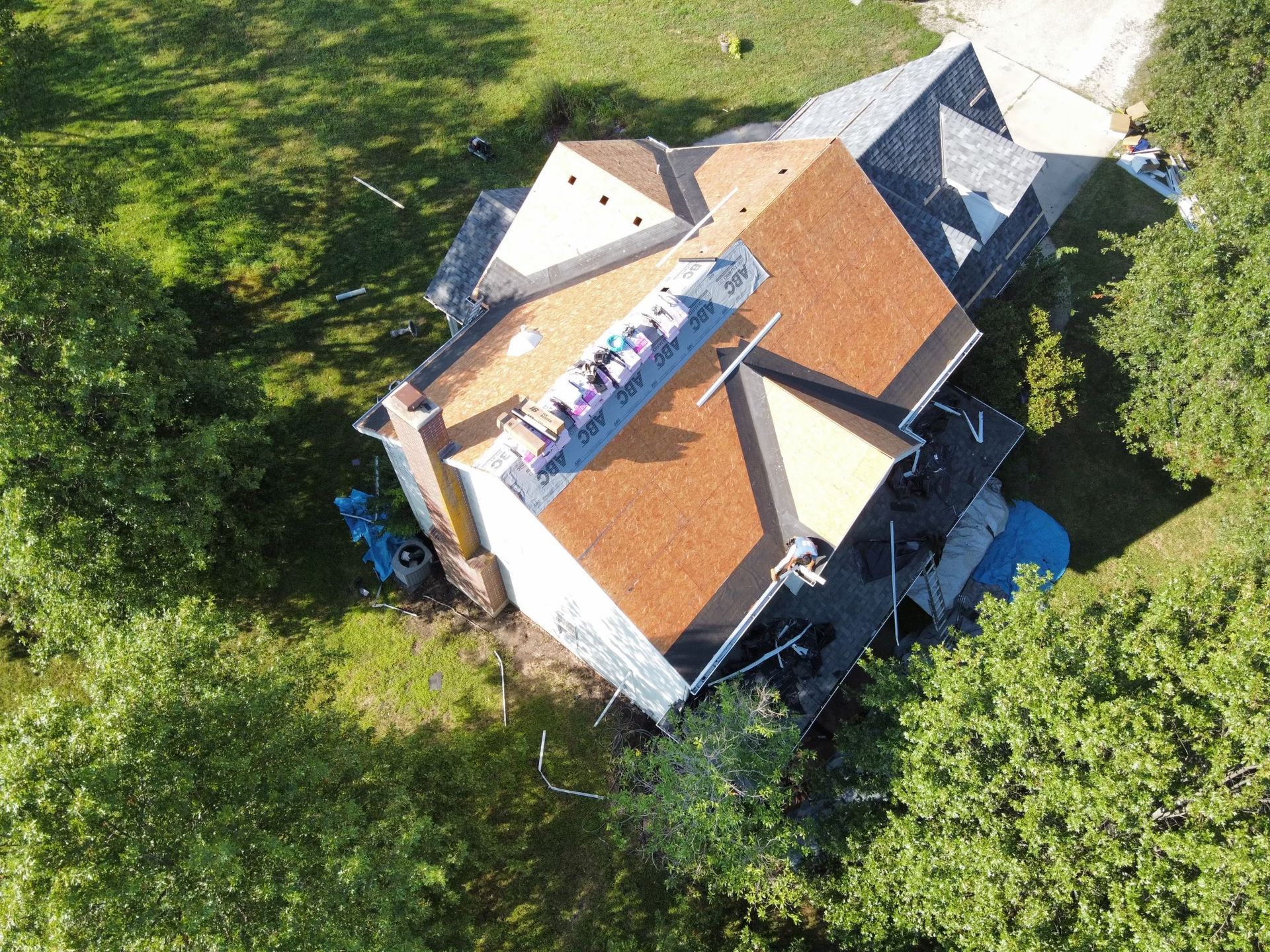 Aerial view of a house with a brown roof, surrounded by green trees and grass, with construction materials visible.