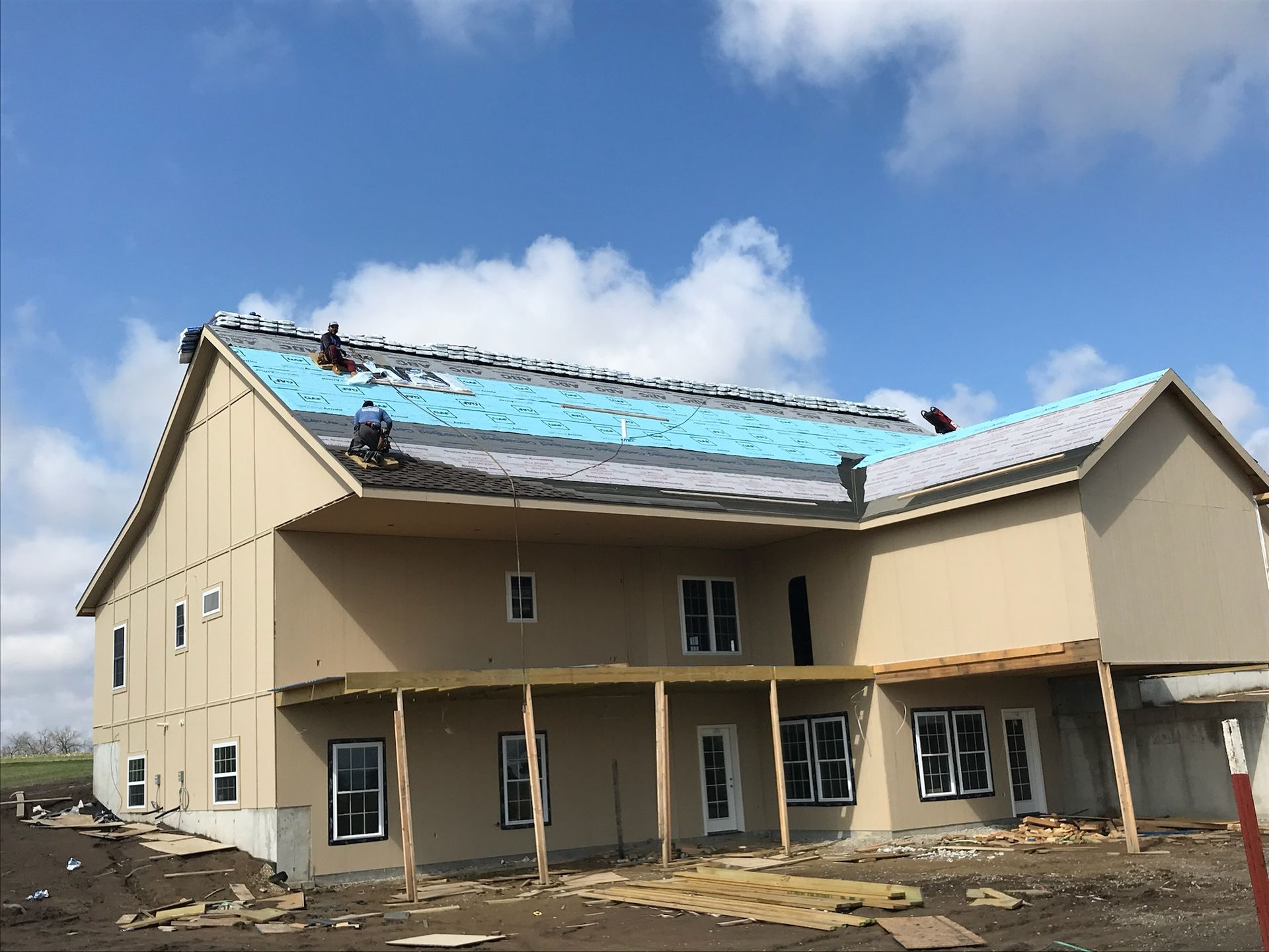 Construction workers on a roof laying shingles on a two-story house under a blue sky.