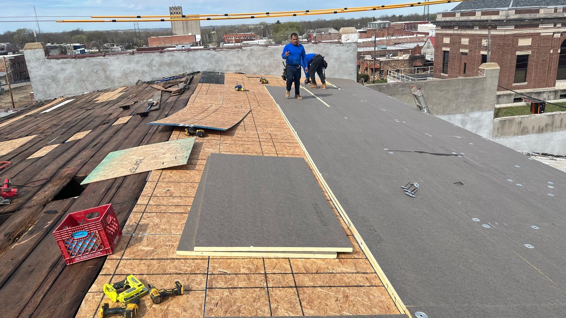 Workers on a rooftop replacing roofing materials. Wooden planks, insulation, and tools are visible.