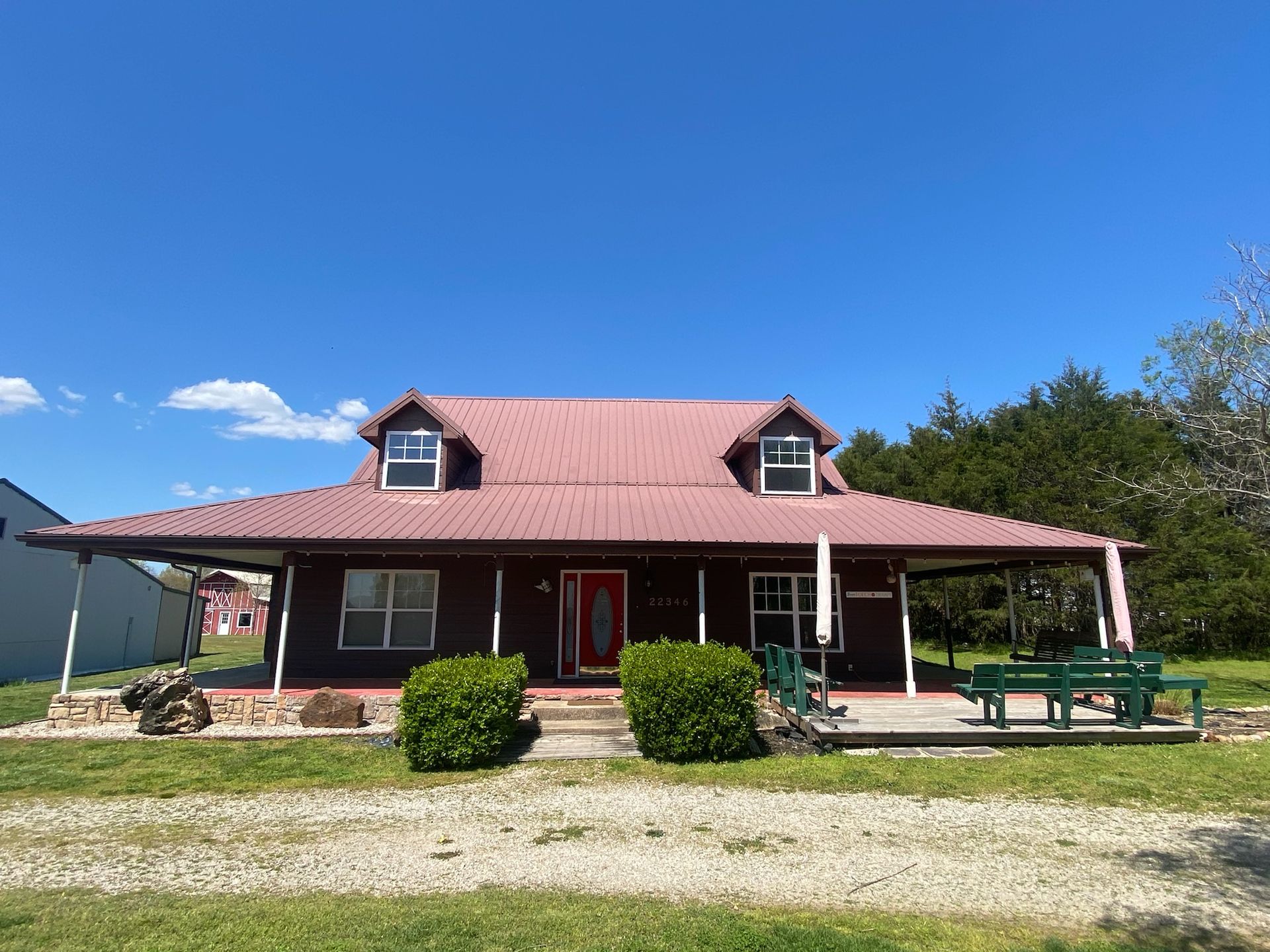 Brown house with red roof, porch, and picnic table on a sunny day.