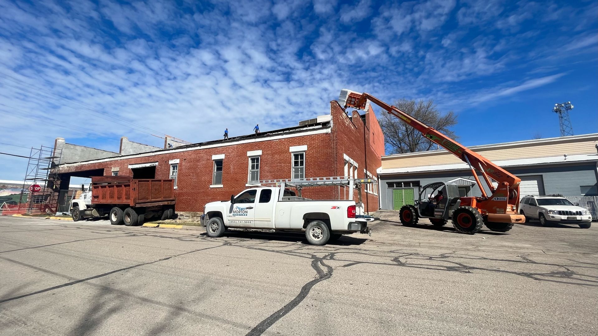 Construction site: red brick building being demolished. Trucks, a crane, and workers are present. Blue sky.