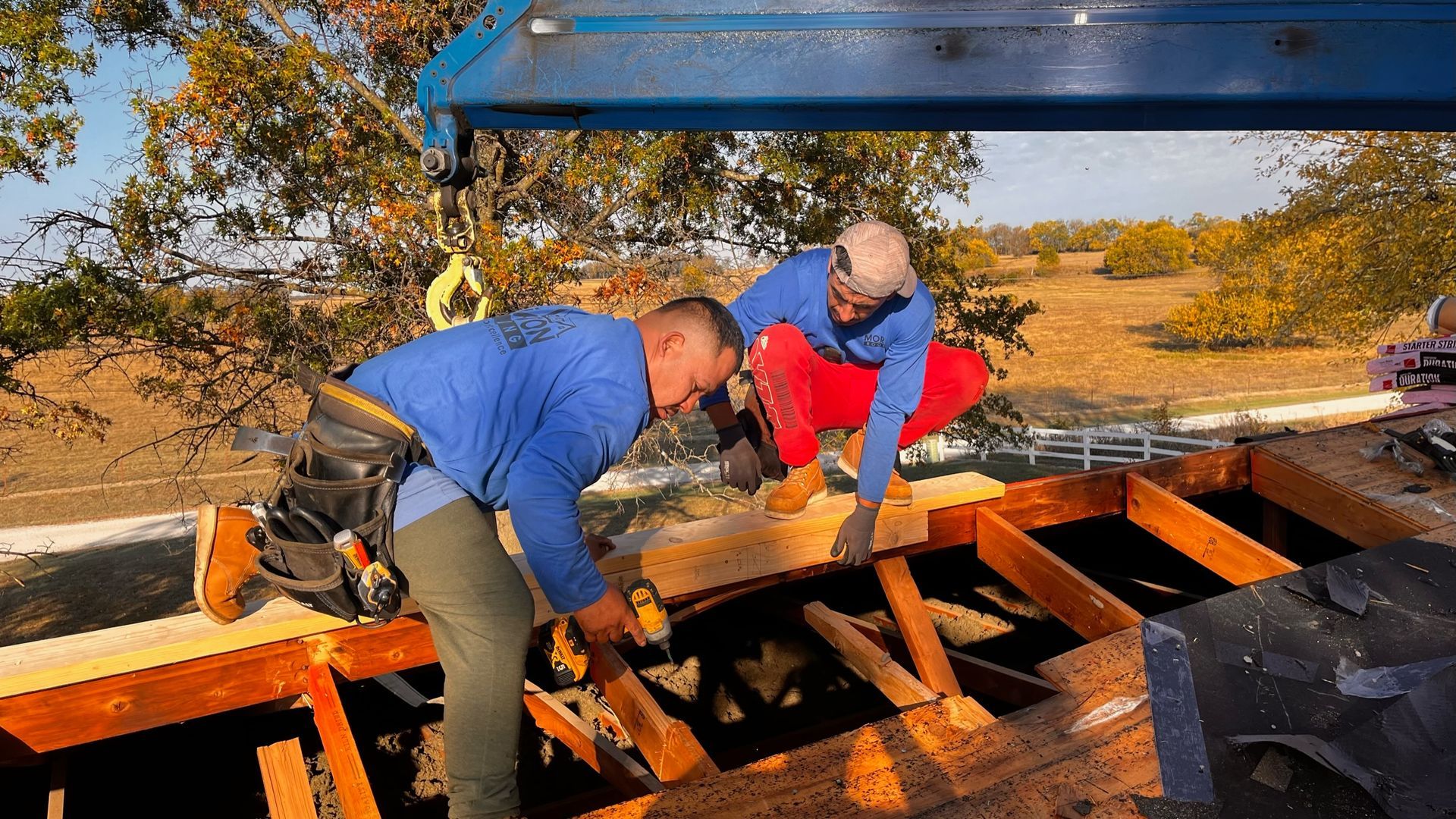 Two construction workers on a roof, one using a drill, the other passing a wood beam. Sunny day.