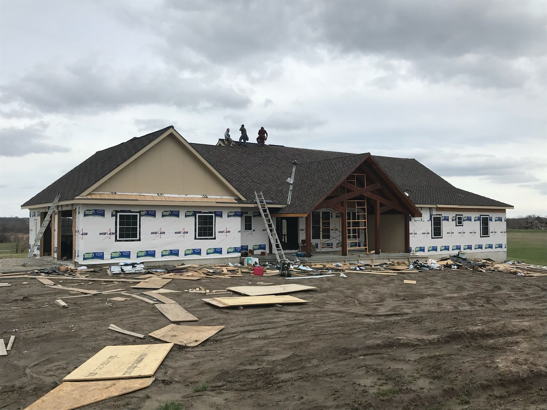 House under construction with roofers on the roof, surrounded by construction materials on a cloudy day.