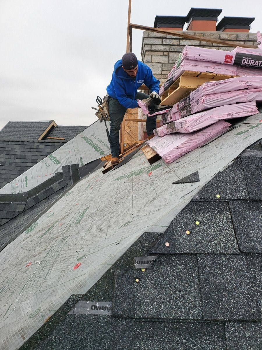 Man on a roof installing insulation and shingles. Gray sky, building materials, and chimney visible.