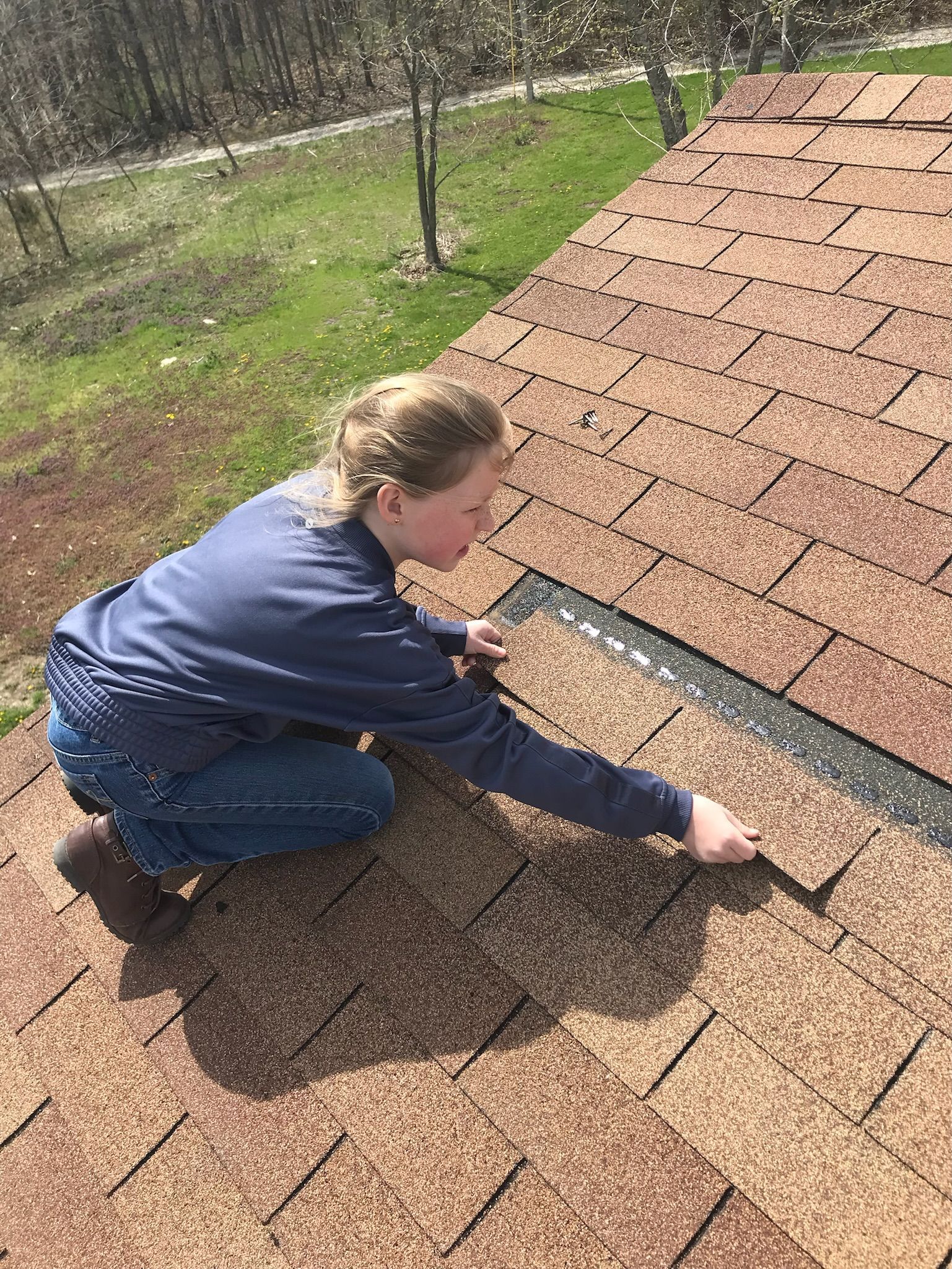 Girl on a brown shingled roof, kneeling, reaching toward a section of missing shingles.