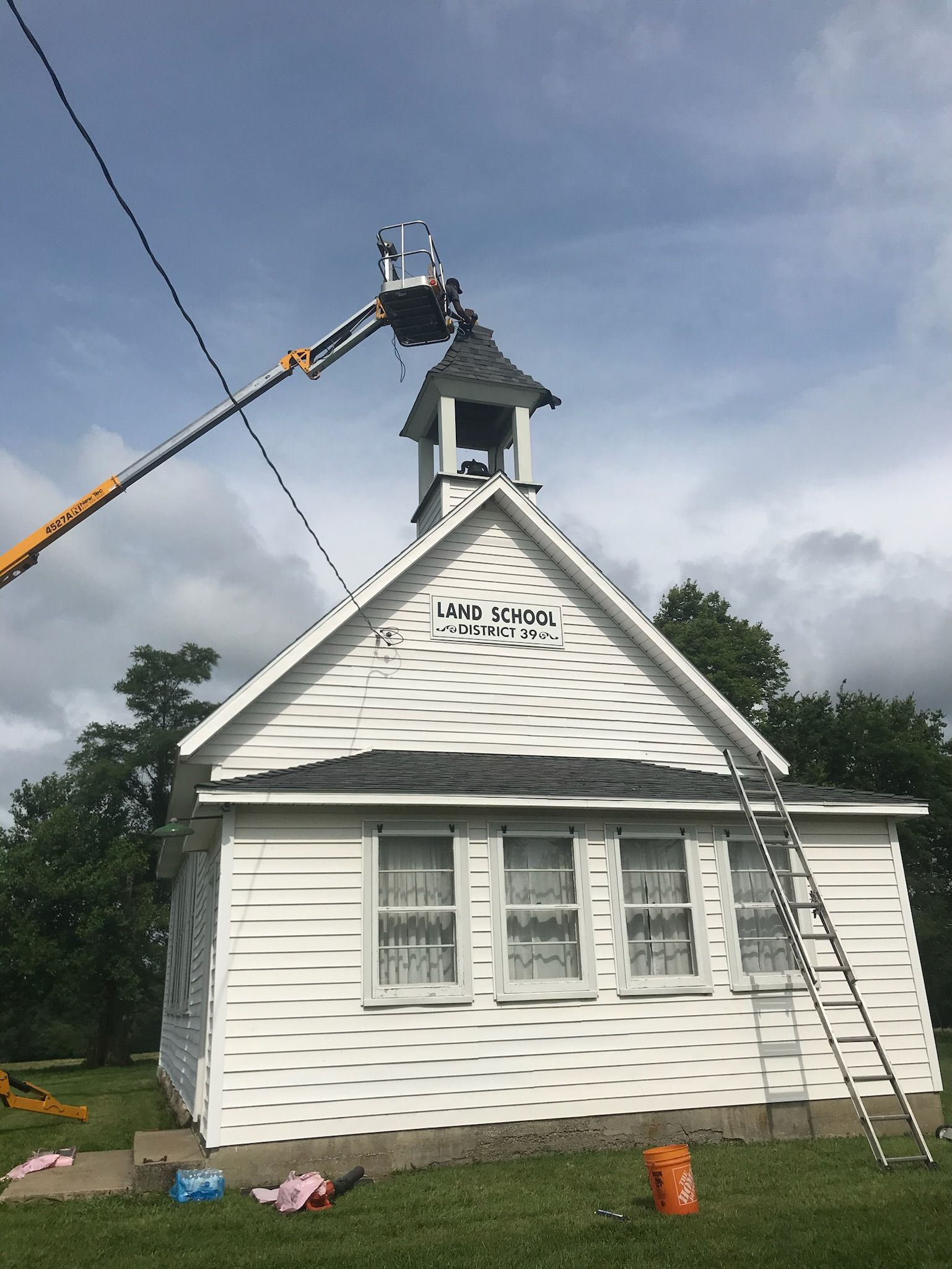 White schoolhouse with people on a lift working on the bell tower under a cloudy sky. A ladder rests against the building.