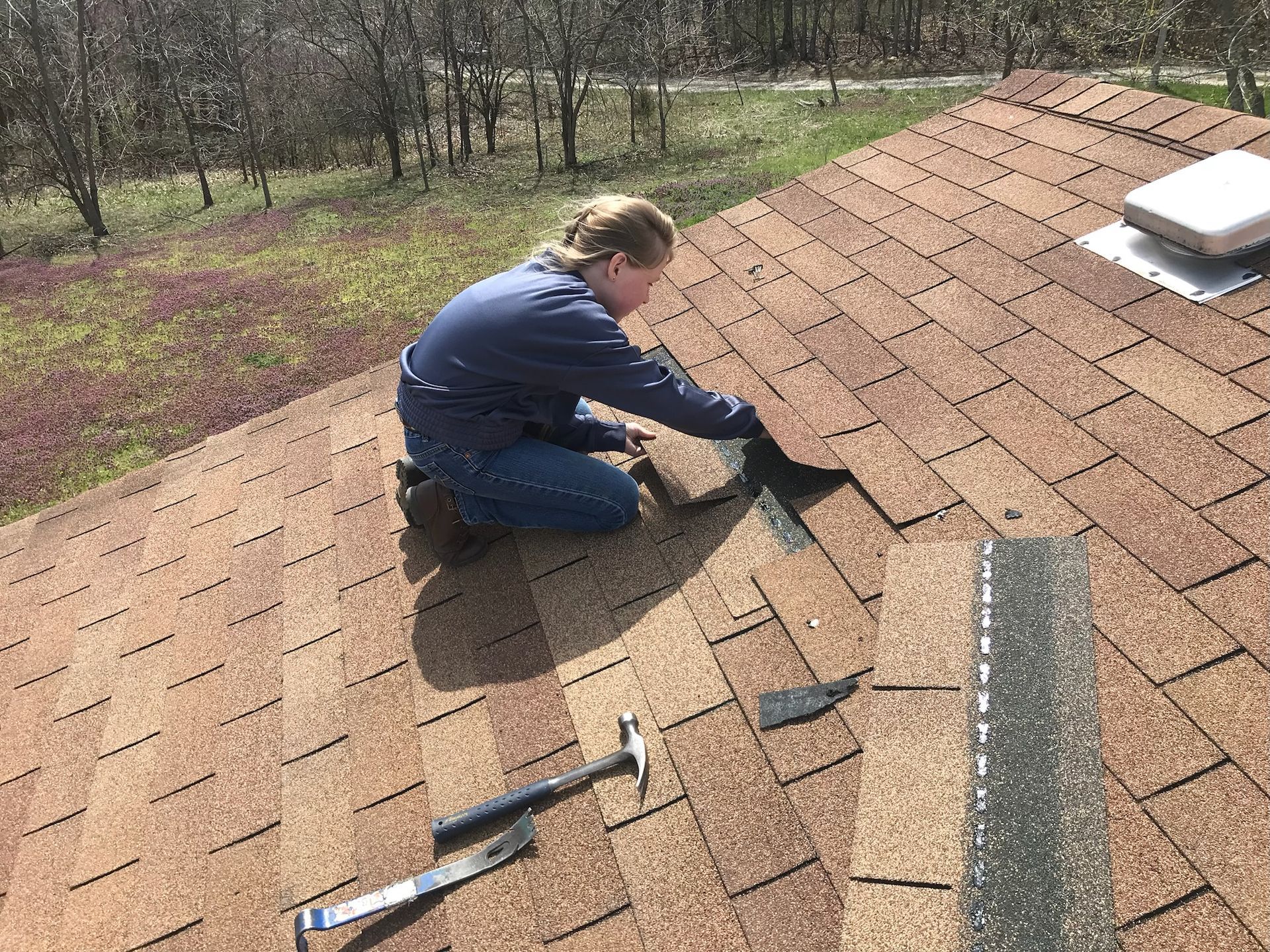 Person on a brown shingled roof, repairing shingles with a hammer and tools; sunny outdoors.