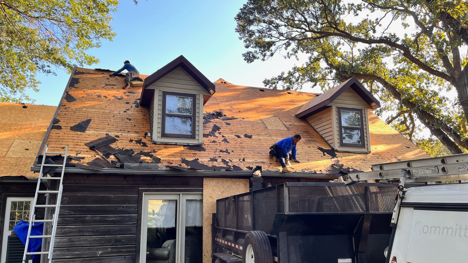Roofers removing shingles from a two-story house with two dormers; a truck bed loaded with debris sits below.
