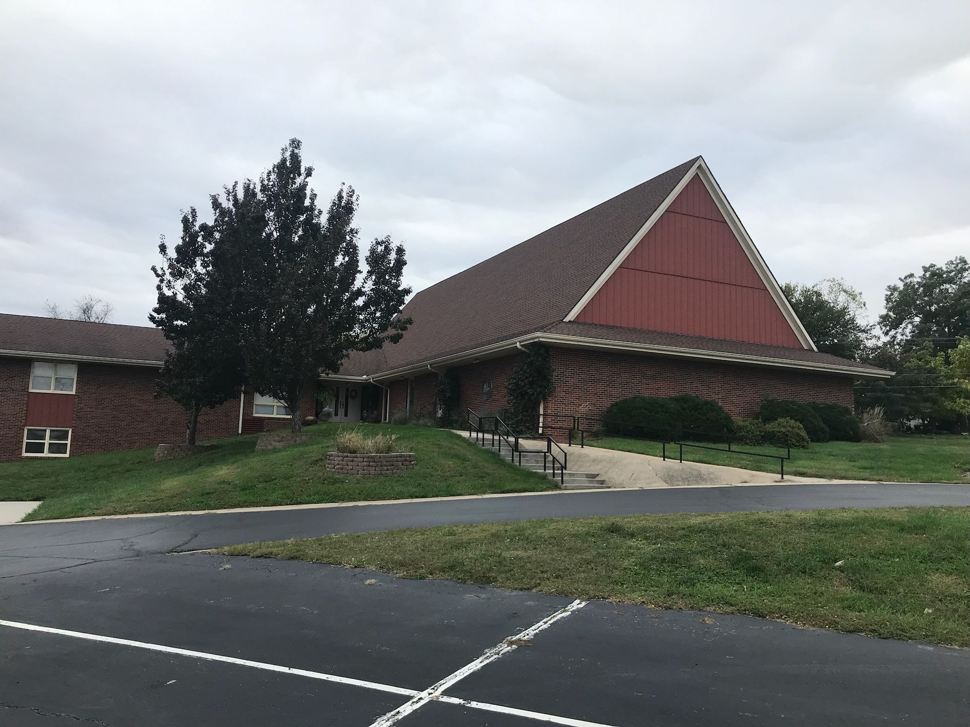 Church building with brown roof and red accent, on a cloudy day. Includes green grass and a parking lot.