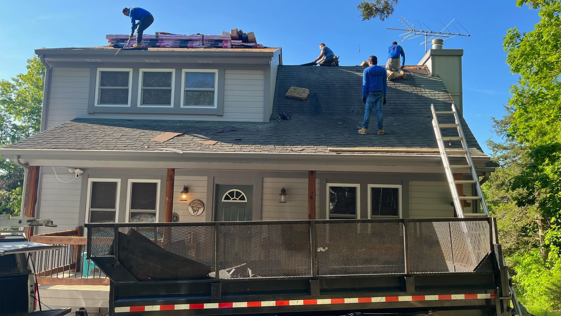 Roofers working on a two-story house with a truck below. Blue shirts, sunny day.