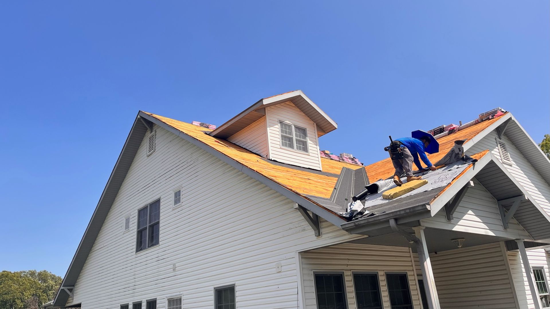 Workers on a roof, replacing shingles. White house, blue sky.