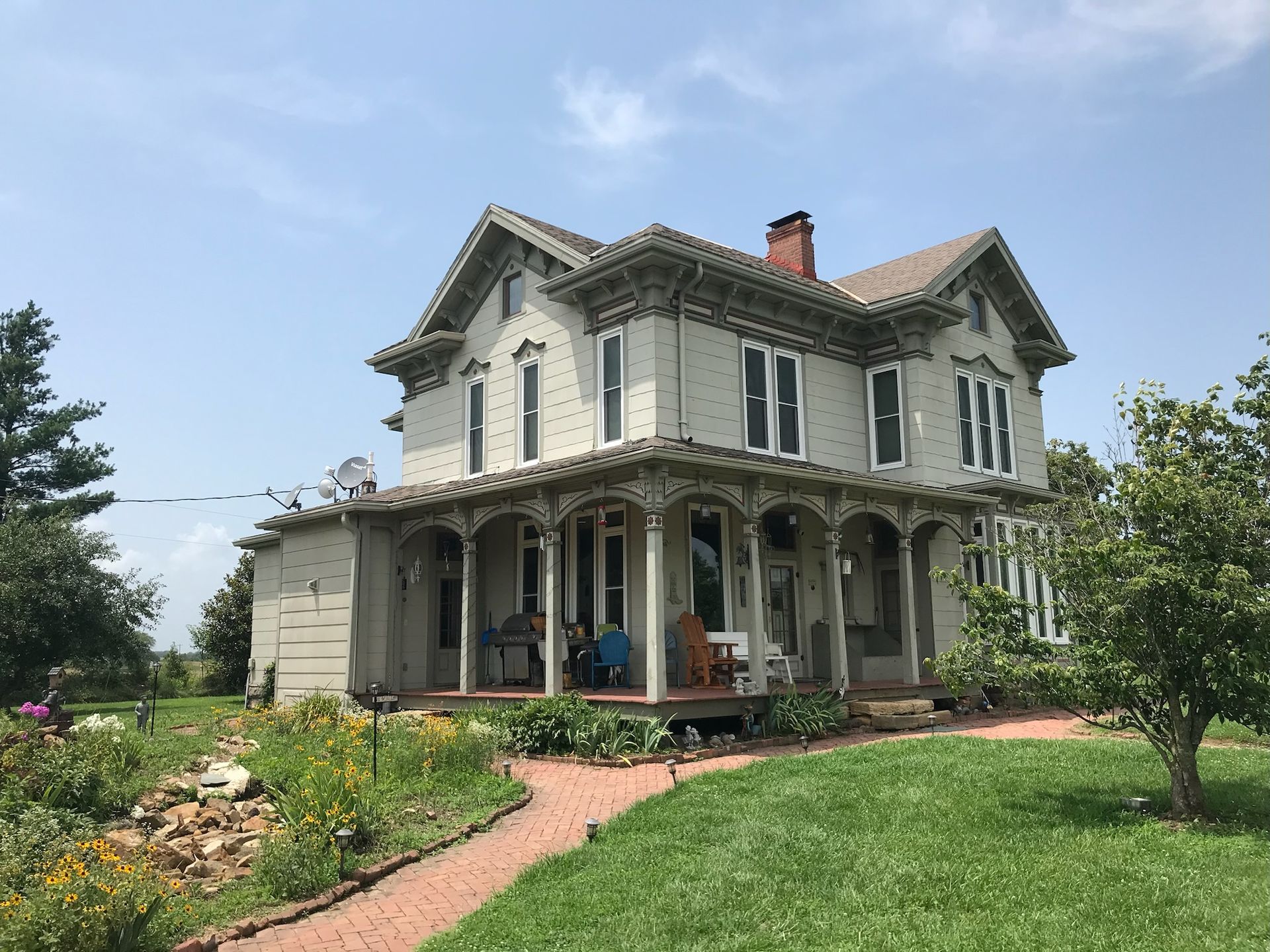 Two-story, light gray Victorian house with a wraparound porch, red brick walkway, and sunny sky.