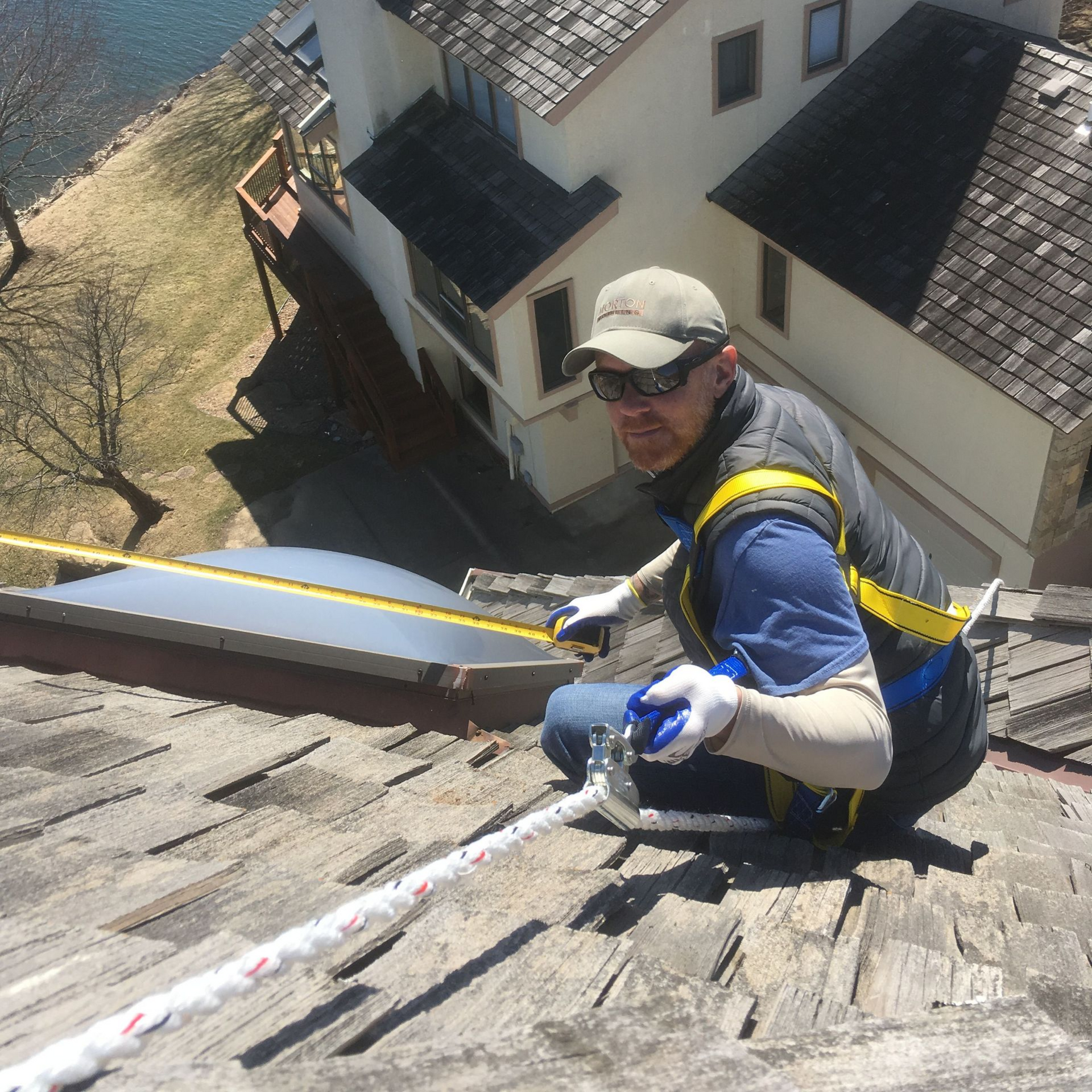 Roofer in safety gear measures on a weathered roof, a house and lake in the background.