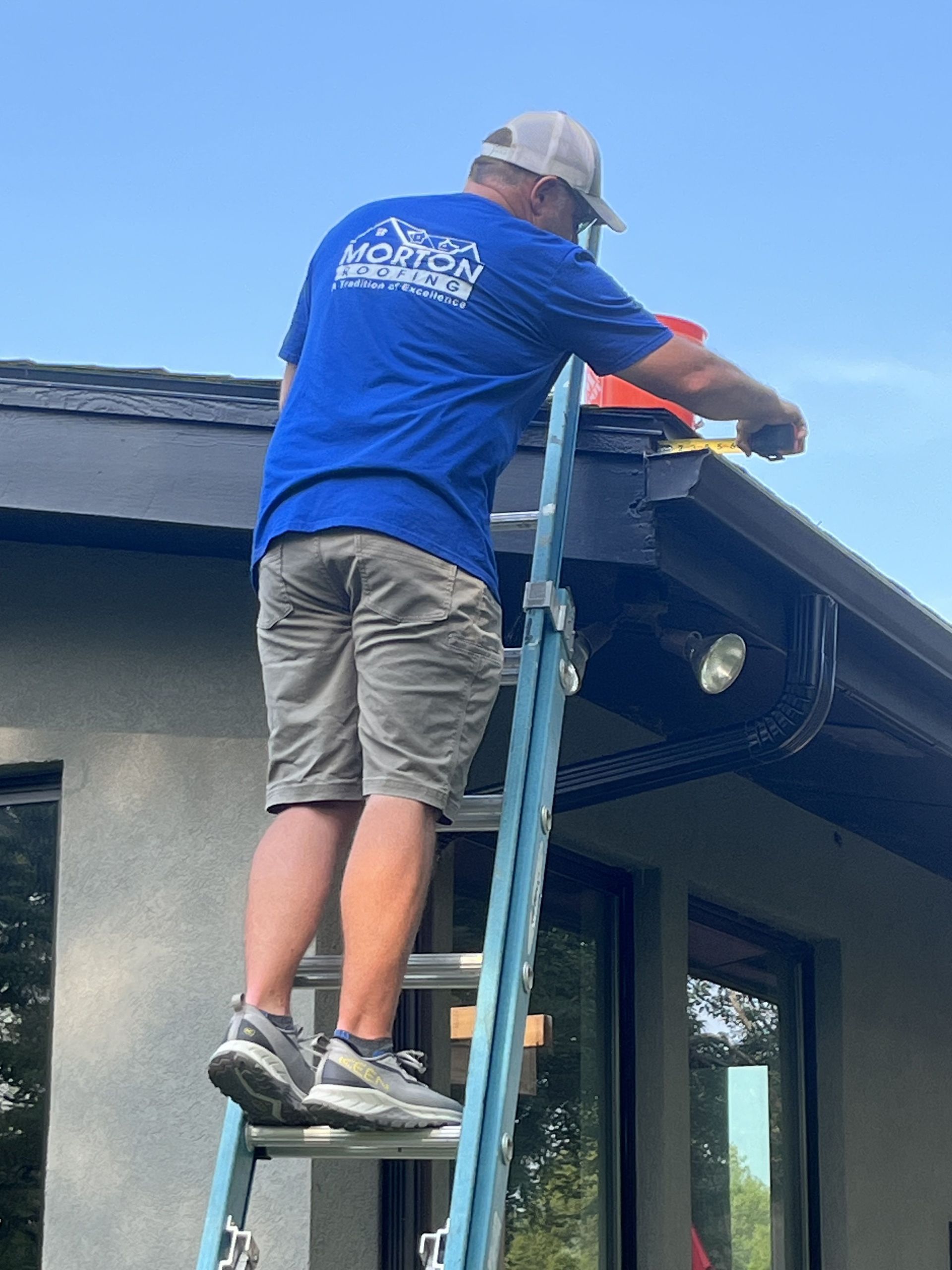 Man on a ladder cleaning a black gutter. He wears a blue shirt, khaki shorts, and a white cap.