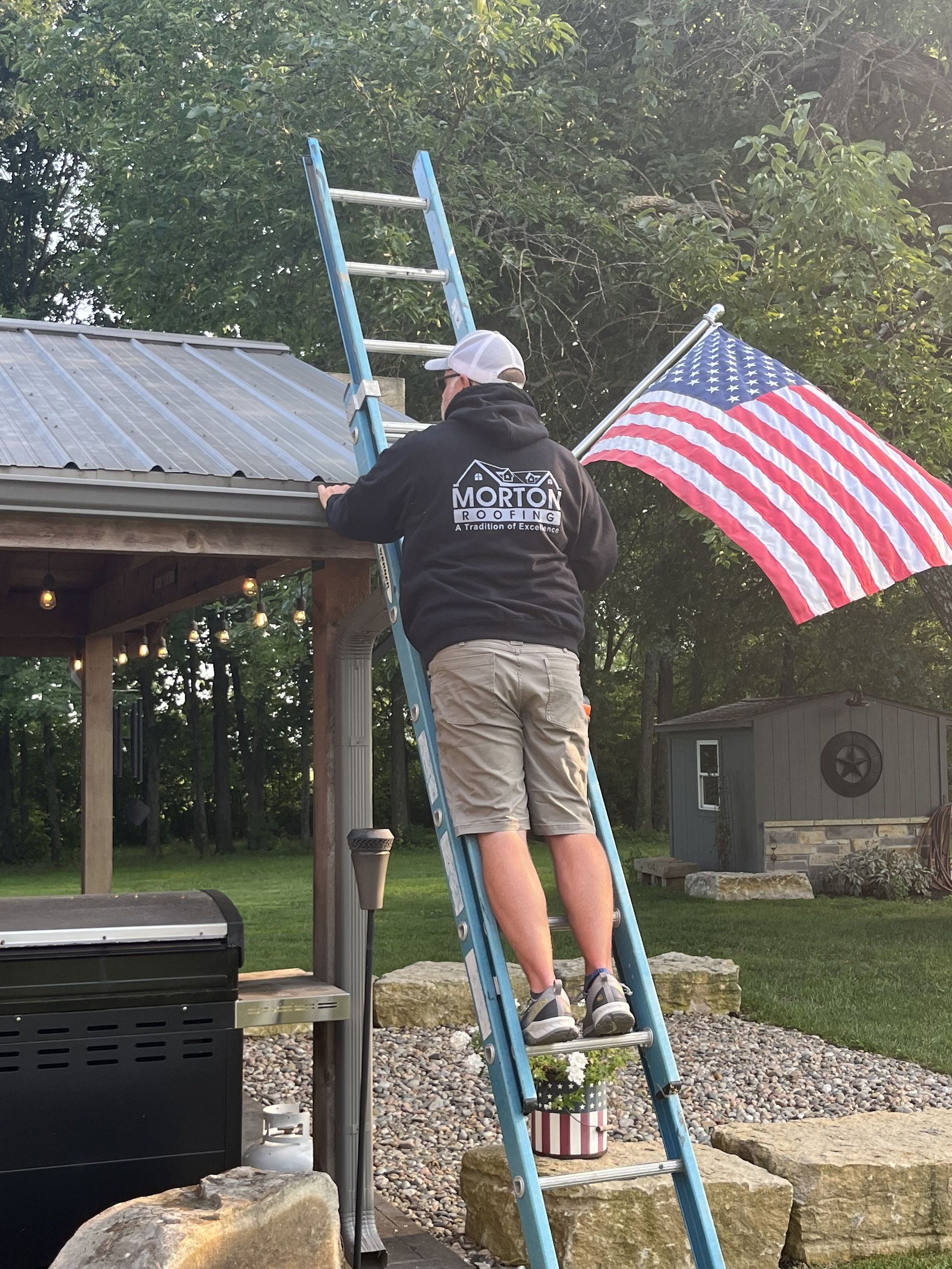 Man on a ladder cleaning a gutter next to an American flag.
