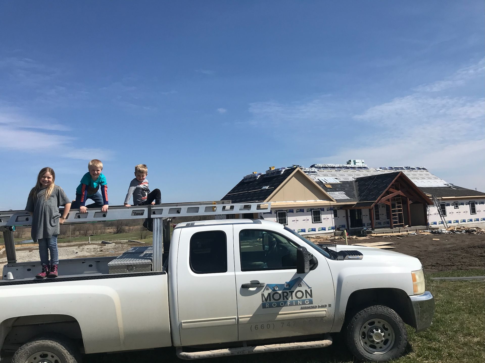 Three children sit on a truck bed in front of a house under construction on a sunny day.
