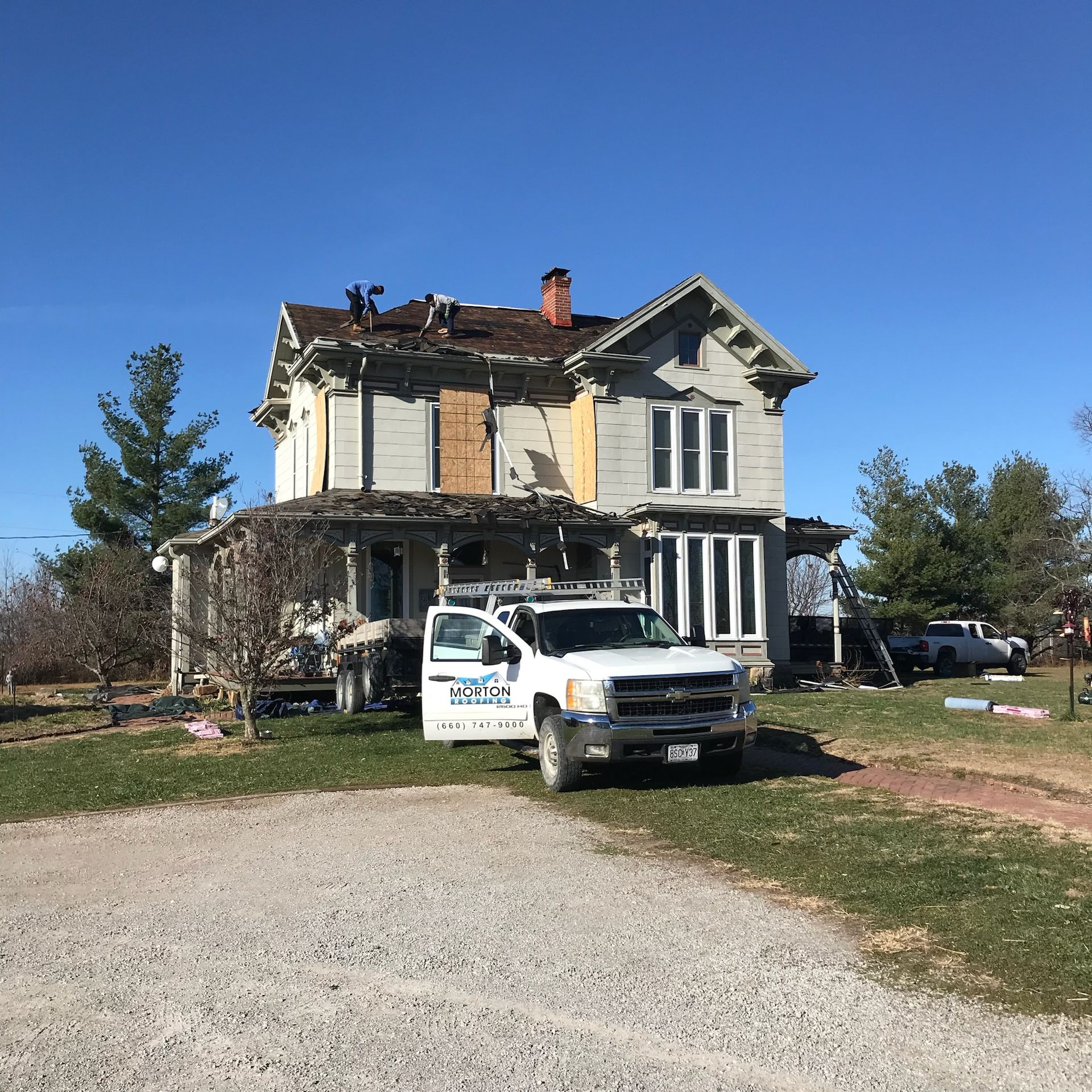 A two-story house being repaired with a white truck in front. Boarded-up windows and a damaged roof.