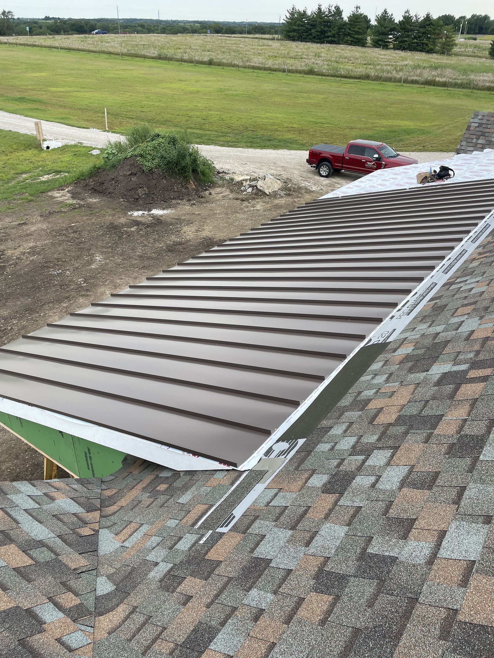 Brown metal roof being installed next to an asphalt shingle roof, with a red truck in the background.