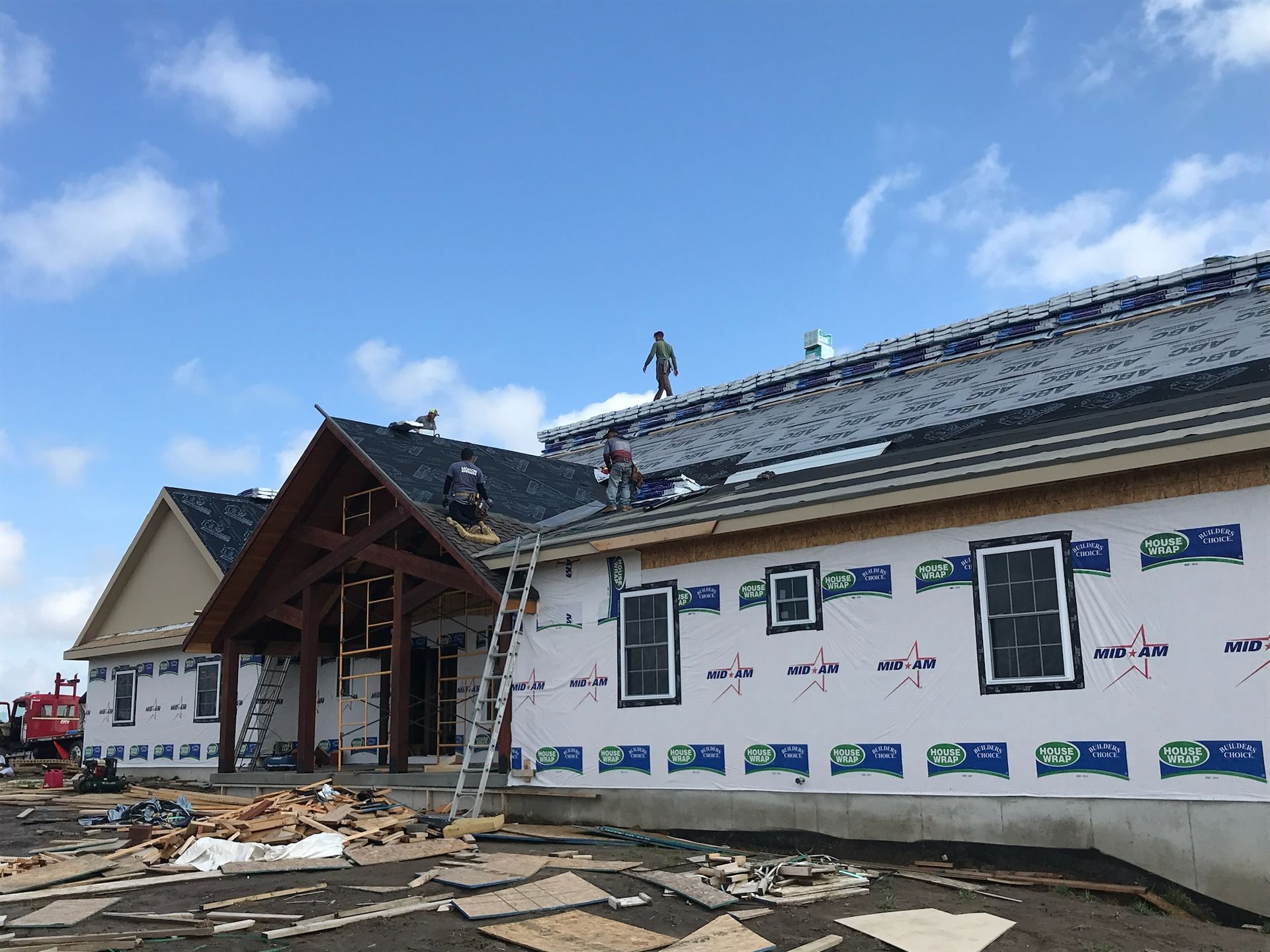 Construction workers installing shingles on a house with a porch, blue sky in the background.