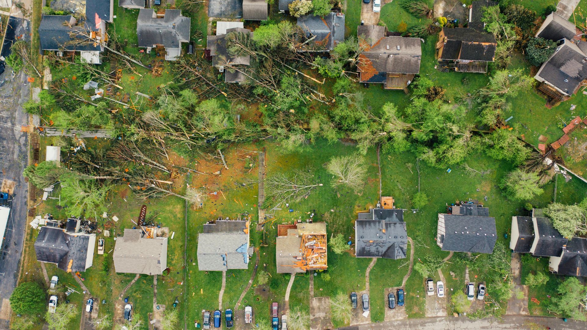 Aerial view of a neighborhood after a storm; damaged houses with missing roofs, fallen trees, and debris on the ground.