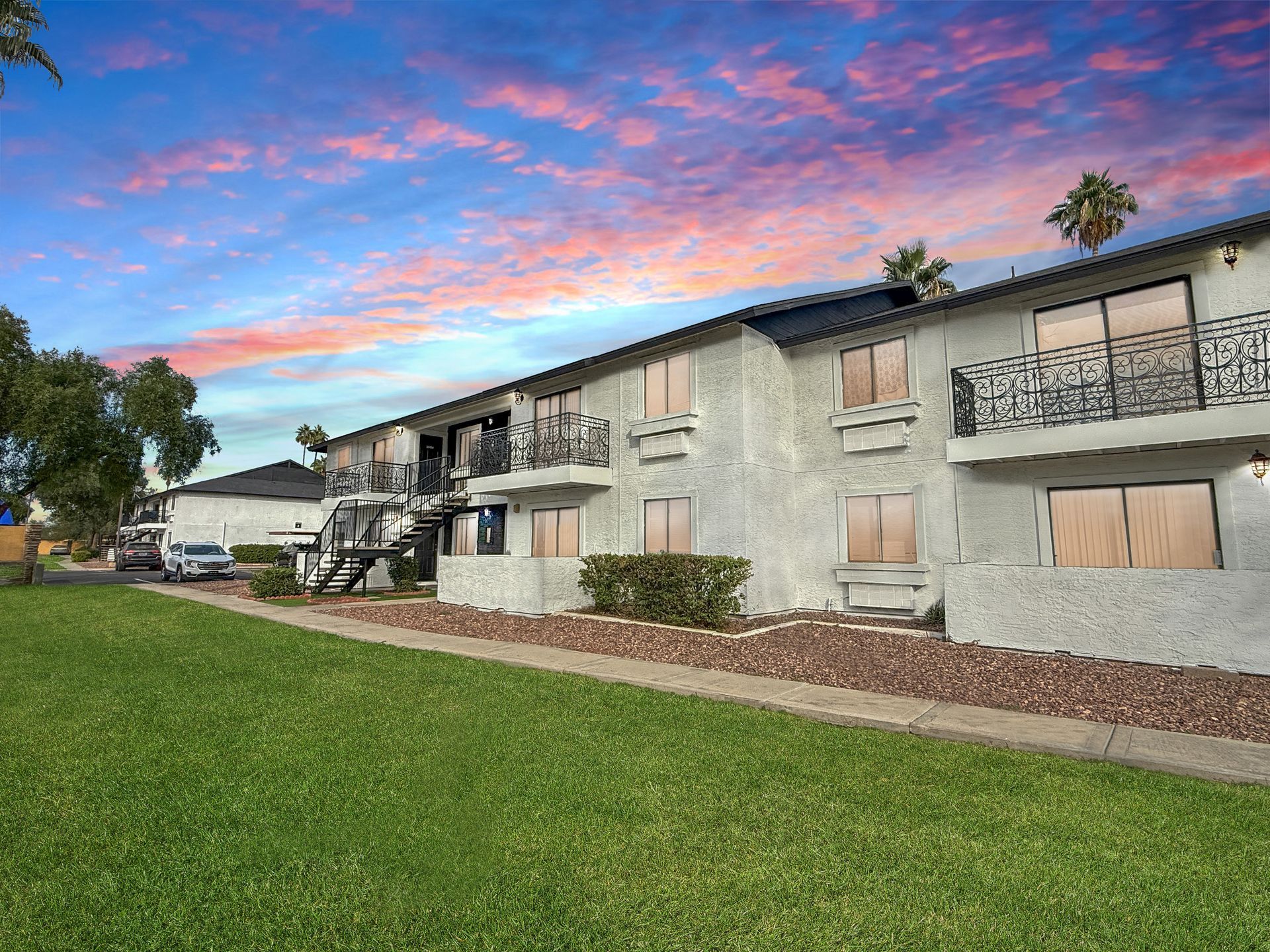 Apartment building with balconies and a green lawn under a colorful sunset sky.