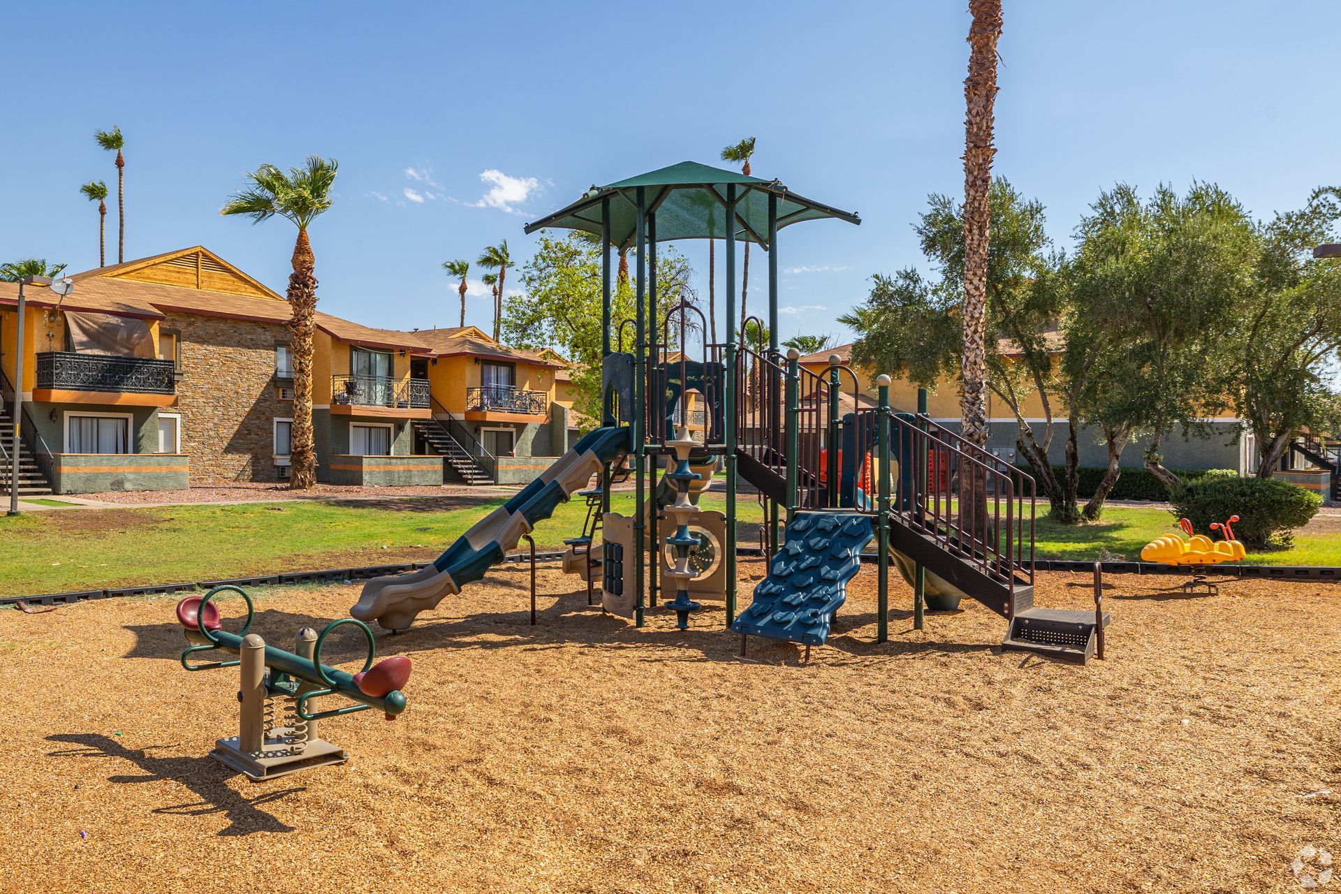 Playground with slides, climbing structures, and a seesaw in a grassy area near buildings.