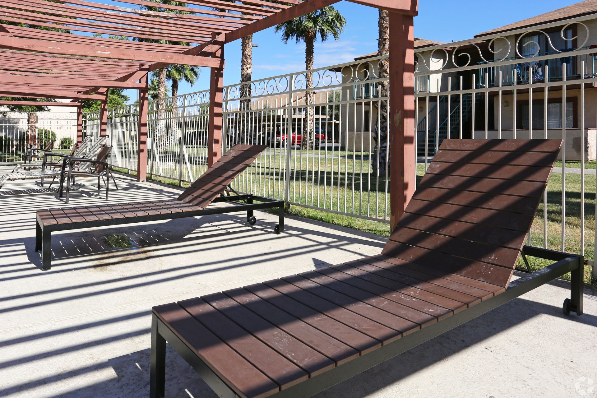Two brown lounge chairs under a pergola, beside a metal fence, next to a building.