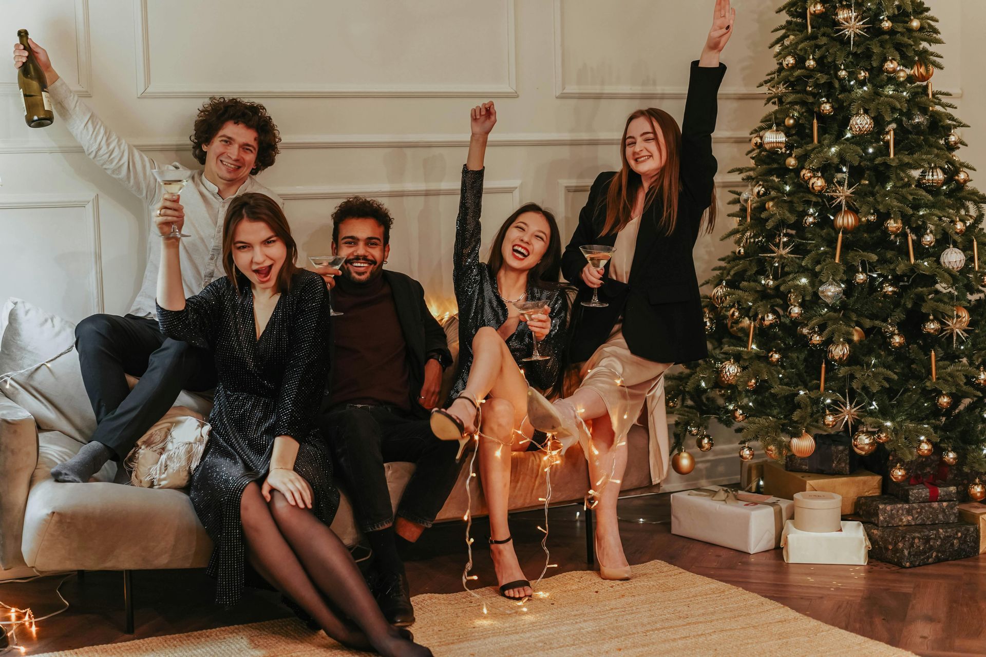 Five people celebrating, sitting on a sofa with Christmas tree. They're smiling, raising glasses and arms. Warm lights.