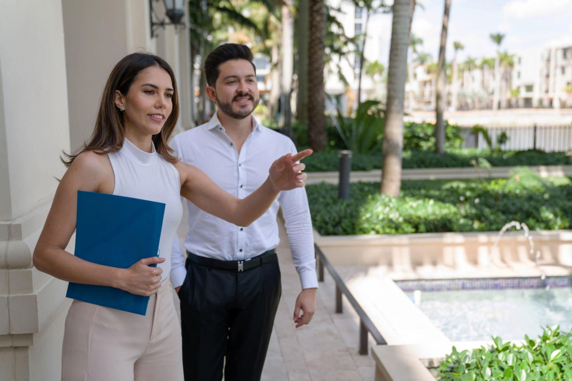 Two professionals discussing real estate, woman pointing, man in white shirt, woman with blue folder, plants in background.