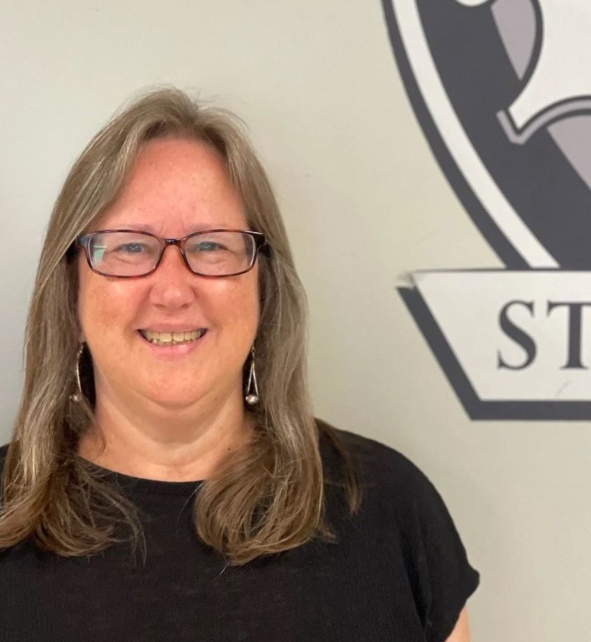 A woman wearing glasses is smiling in front of a sign that says st.