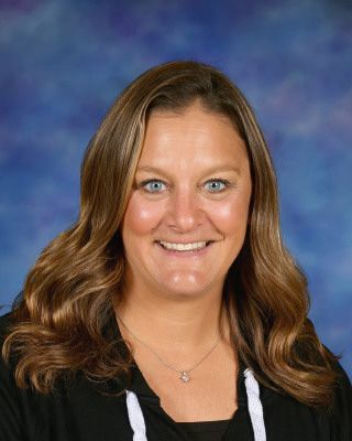 A woman wearing glasses and a necklace is smiling in front of a building.